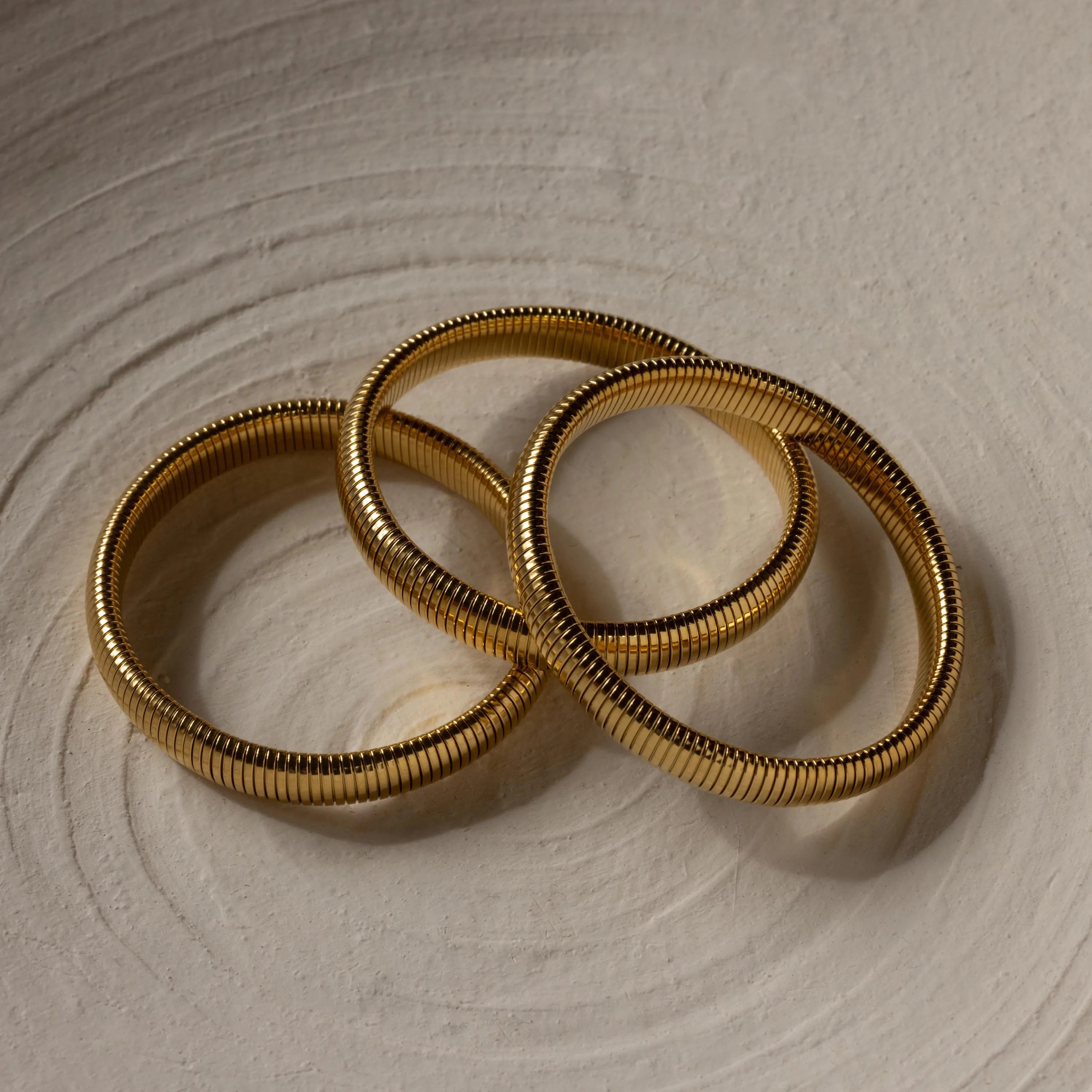 Three Thick Snake Chain Bangles with a ribbed, textured design are elegantly arranged on a light, textured surface.
