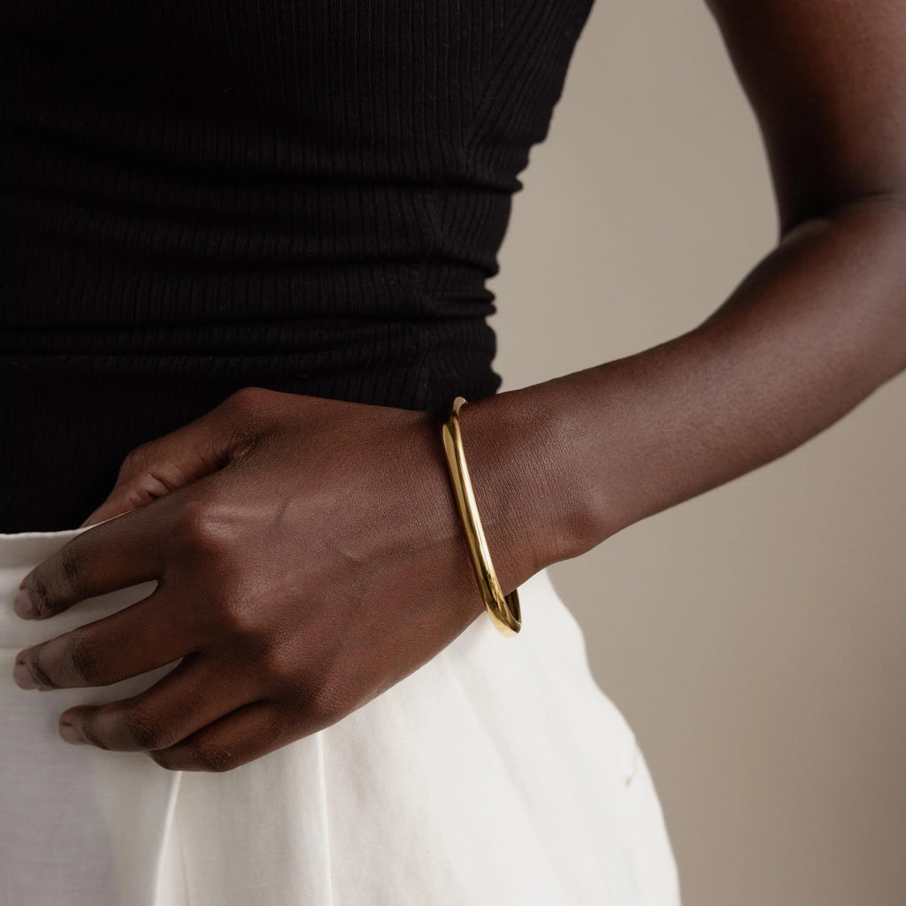 A person wearing the Square Bangle, a statement gold jewelry piece, paired with a black top and white skirt, stands with their hand on their hip.