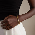 A person wearing the Square Bangle, a statement gold jewelry piece, paired with a black top and white skirt, stands with their hand on their hip.