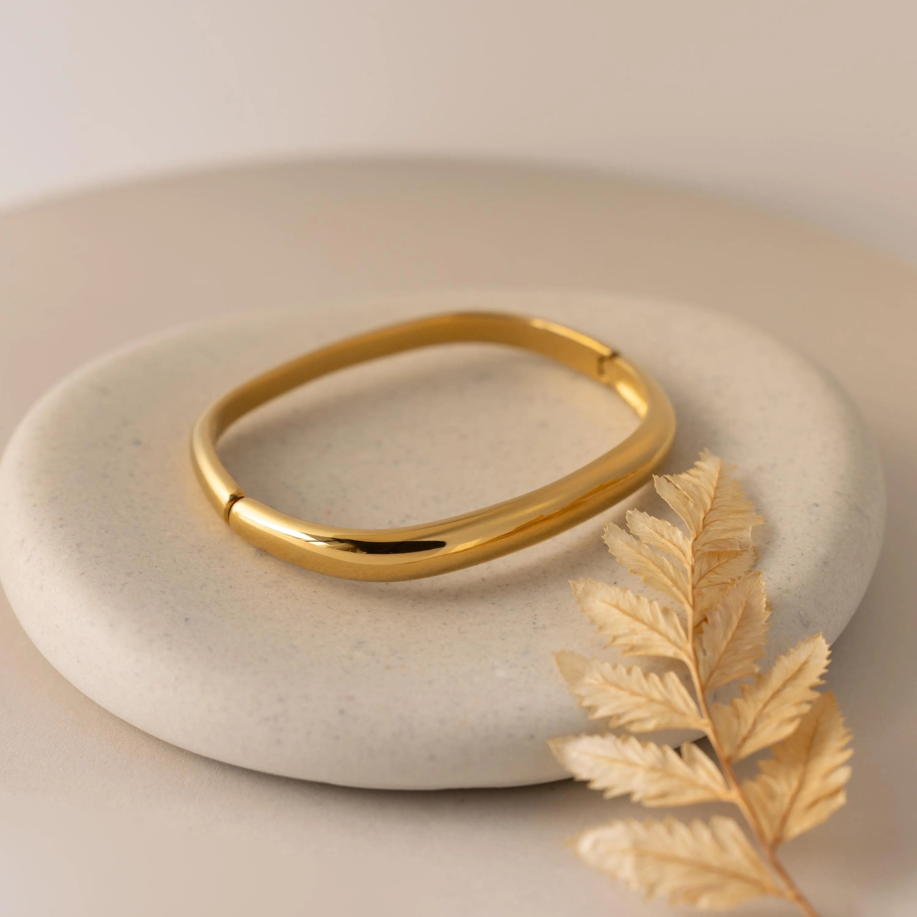 The Square Bangle in gold sits atop a round beige stone, accompanied by a dried golden leaf.