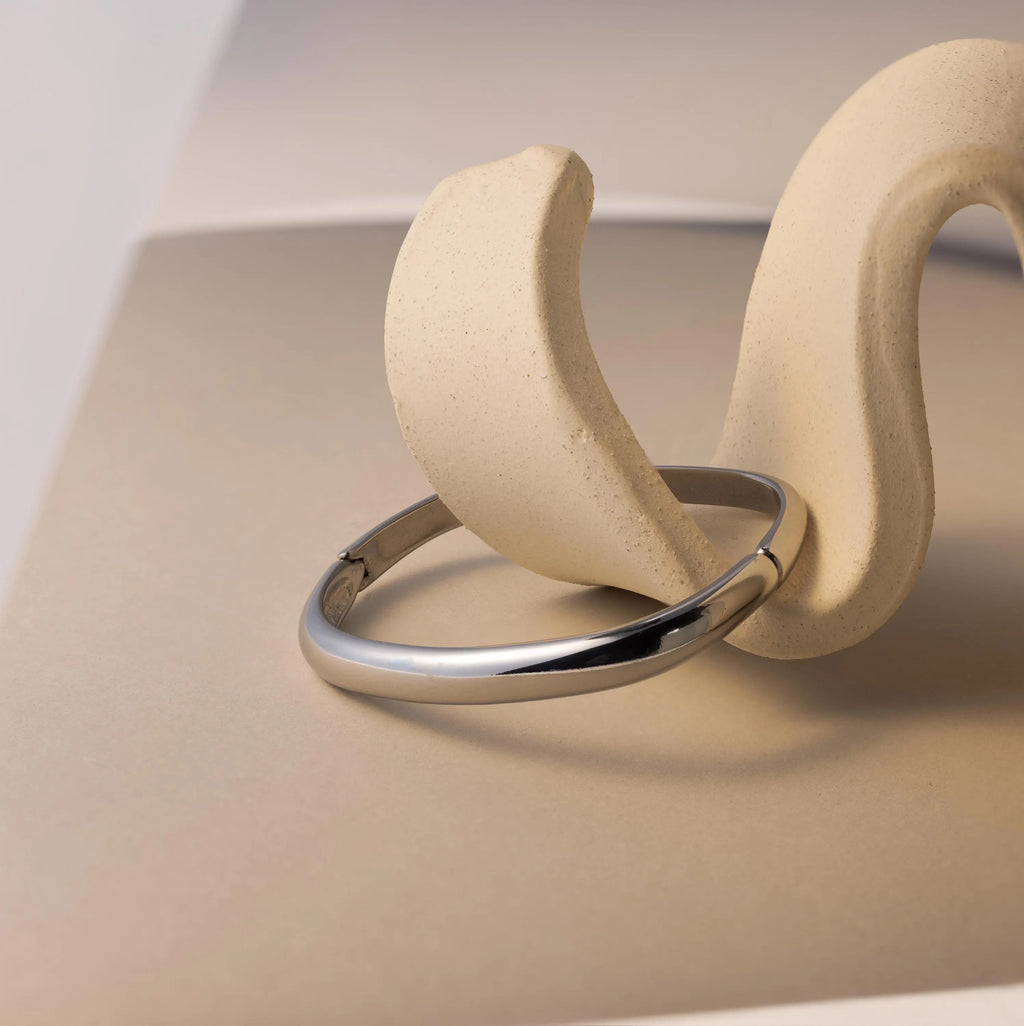 The Square Bangle, a geometric silver bracelet, is showcased on a curved beige sculptural stand against a neutral backdrop, highlighting its striking statement style.