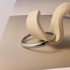 The Square Bangle, a geometric silver bracelet, is showcased on a curved beige sculptural stand against a neutral backdrop, highlighting its striking statement style.
