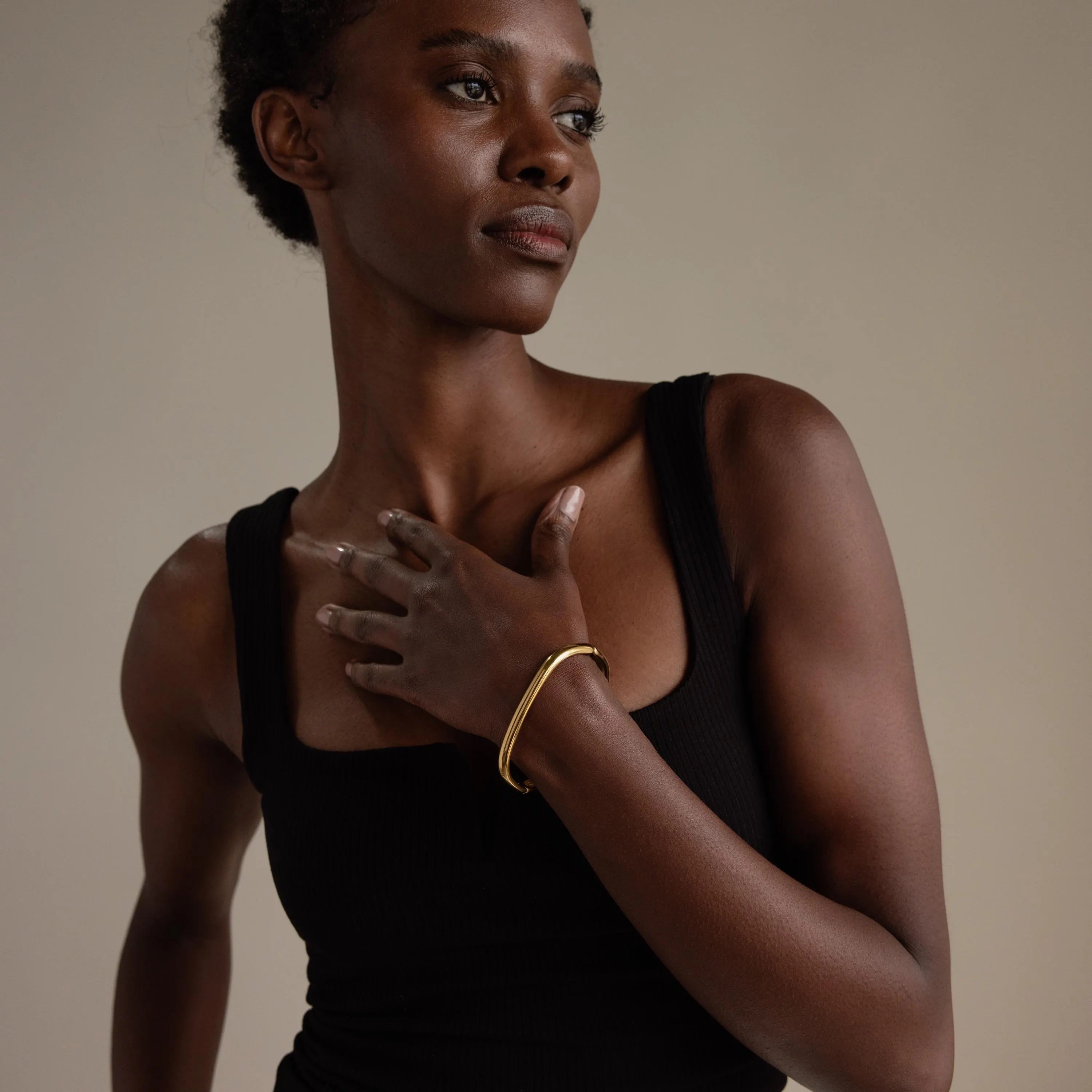 A woman in a black tank top gazes to the side with her hand on her chest, wearing the simple Square Bangle.