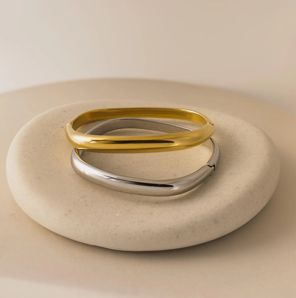 A gold and a silver Square Bangle are displayed on a smooth, round beige stone against a neutral background.