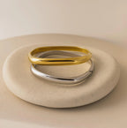 A gold and a silver Square Bangle are displayed on a smooth, round beige stone against a neutral background.