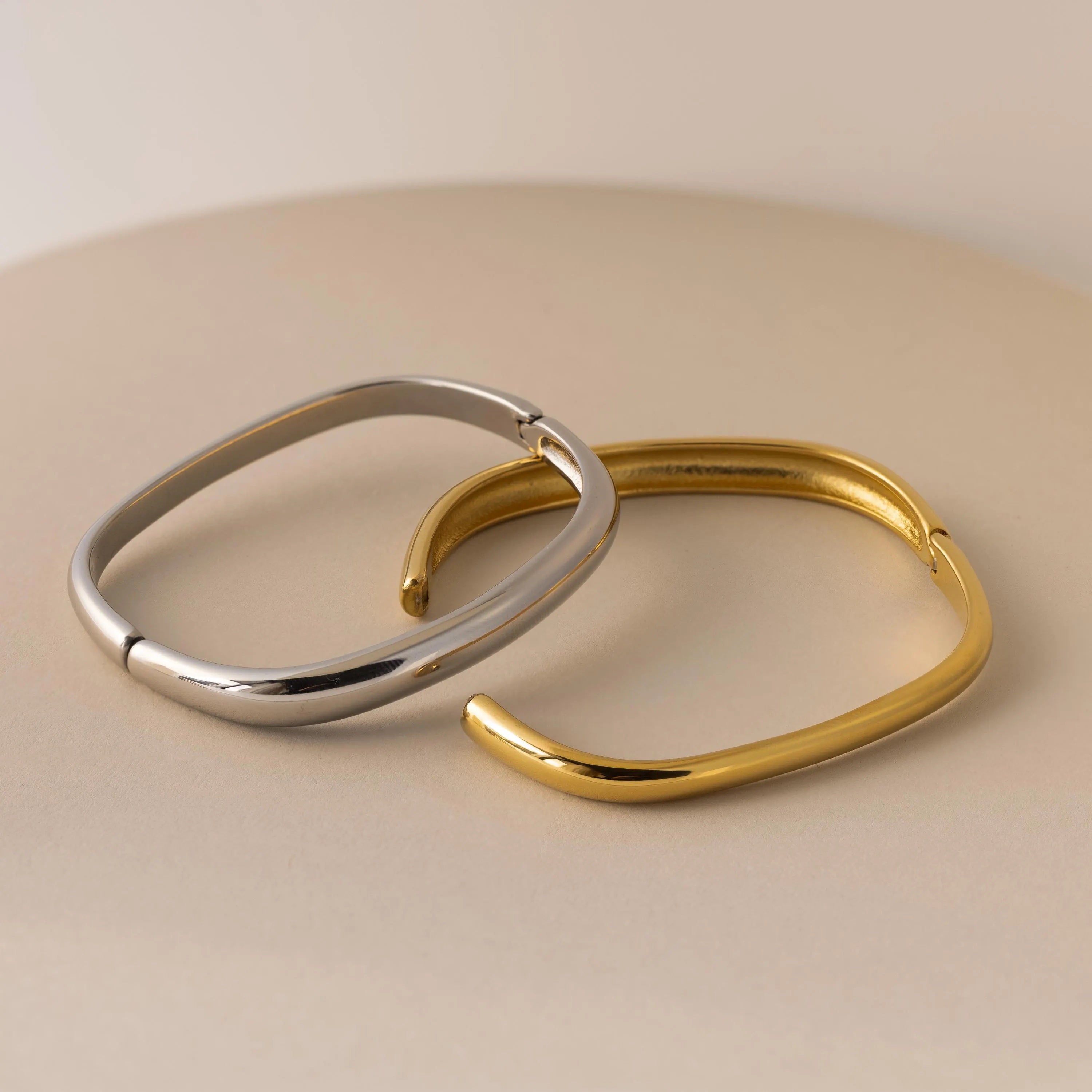 The Square Bangle features two open, square-shaped bangles in silver and gold, displayed on a beige surface—ideal statement jewelry for anyone who loves geometric bracelet styles.