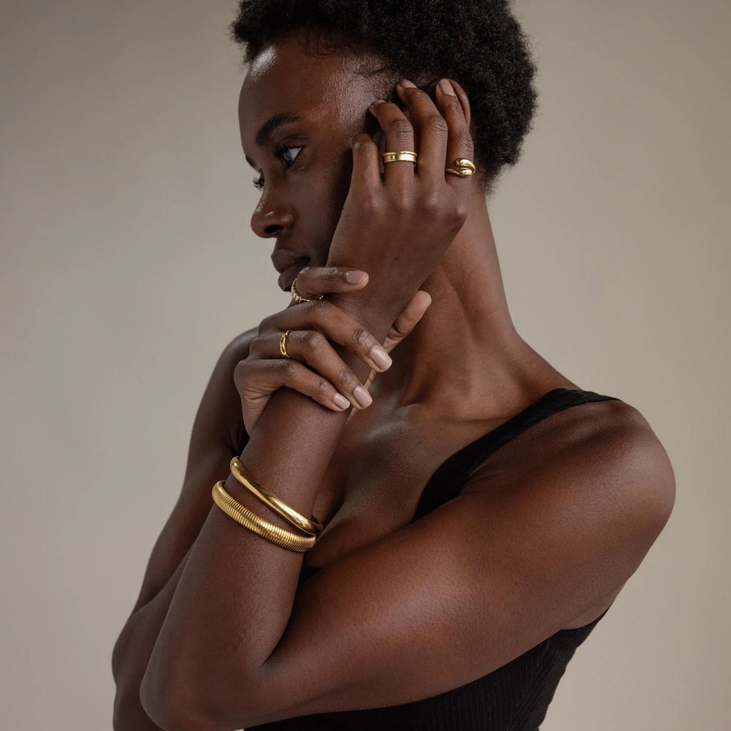 A woman in a black top showcases the Square Bangle along with gold rings and bracelets, posing with her hands near her face against a neutral background.