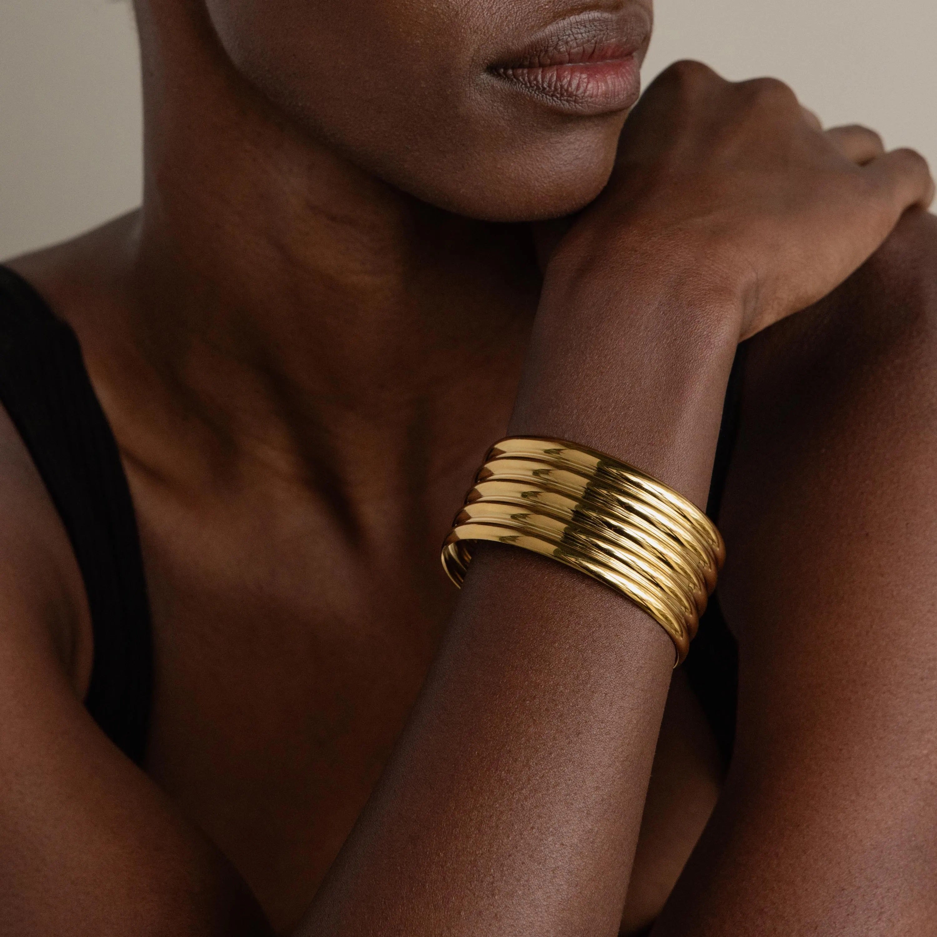 A woman in a black top wears a stack of shiny gold bangles and the Ribbed Cuff Bracelet with ridge detailing on her wrist, set against a neutral background.
