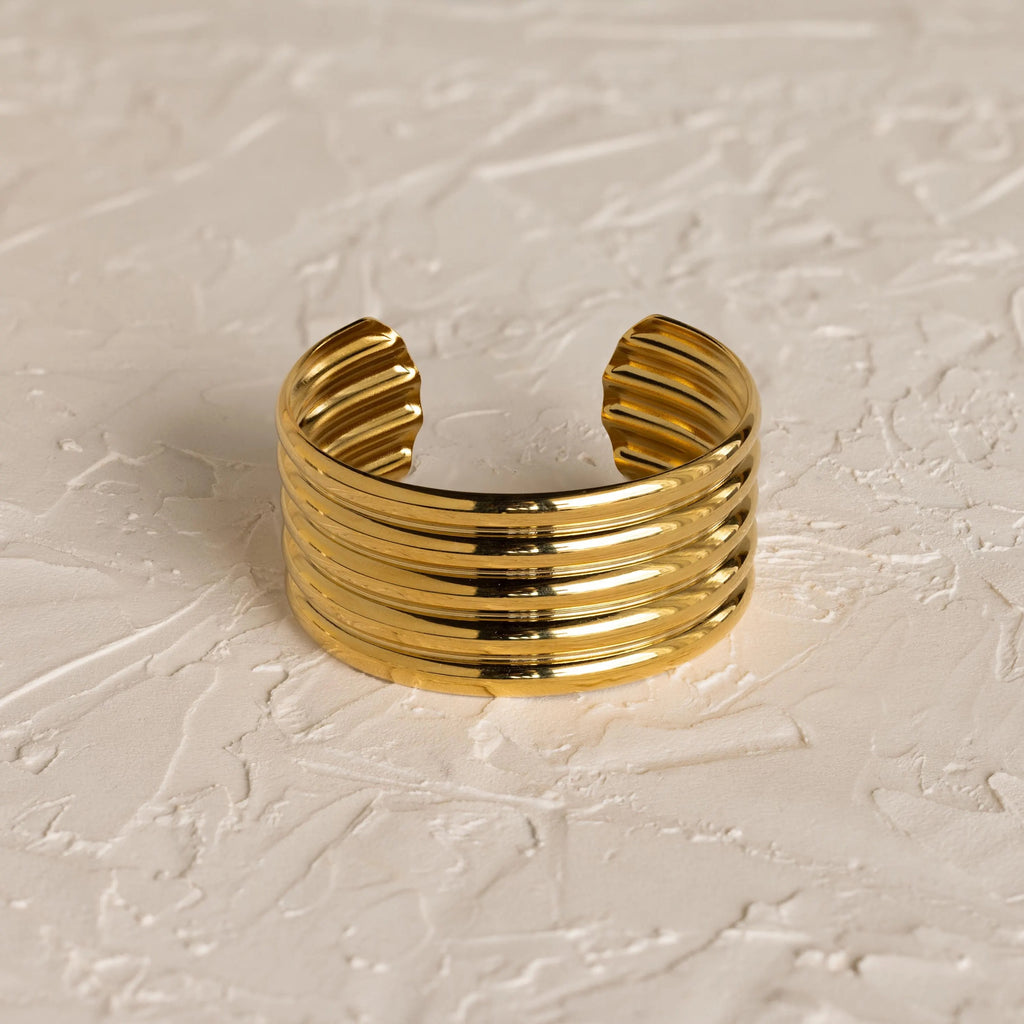 The Ribbed Cuff Bracelet features wide gold ridges and detailed texture, displayed on a textured off-white surface.