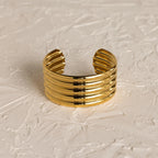 The Ribbed Cuff Bracelet features wide gold ridges and detailed texture, displayed on a textured off-white surface.