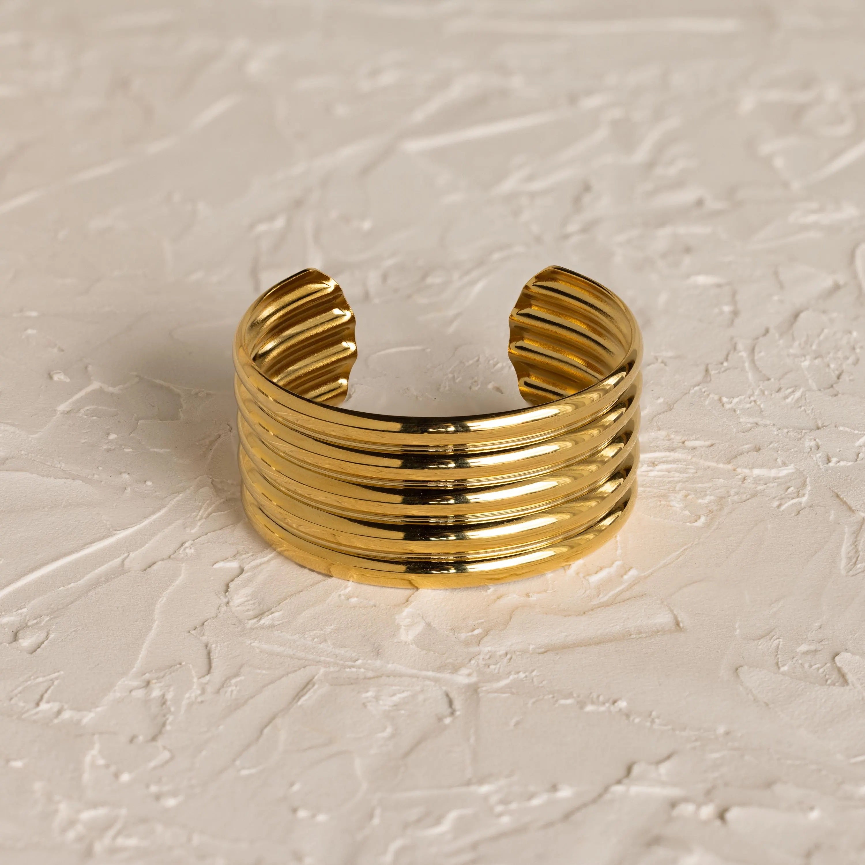 The Ribbed Cuff Bracelet features wide gold ridges and detailed texture, displayed on a textured off-white surface.