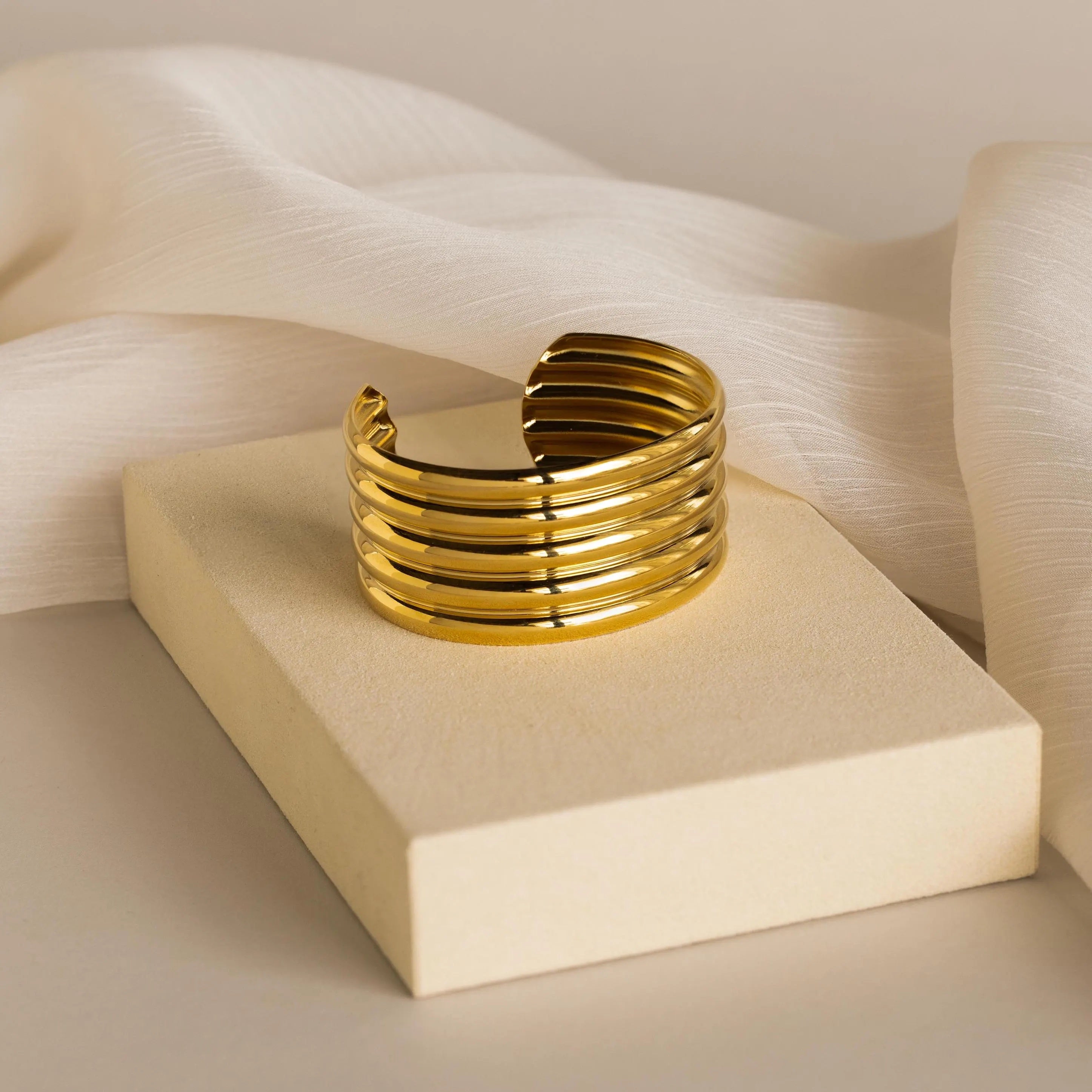 The Ribbed Cuff Bracelet features elegant ridge detailing and is showcased on a cream block against a soft, neutral fabric background.