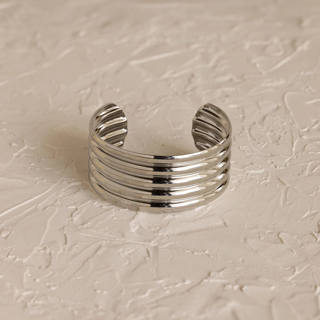 The Ribbed Cuff Bracelet is a wide silver sculpted cuff with five raised horizontal ridges, shown on a textured light-colored surface.