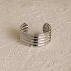 The Ribbed Cuff Bracelet is a wide silver sculpted cuff with five raised horizontal ridges, shown on a textured light-colored surface.