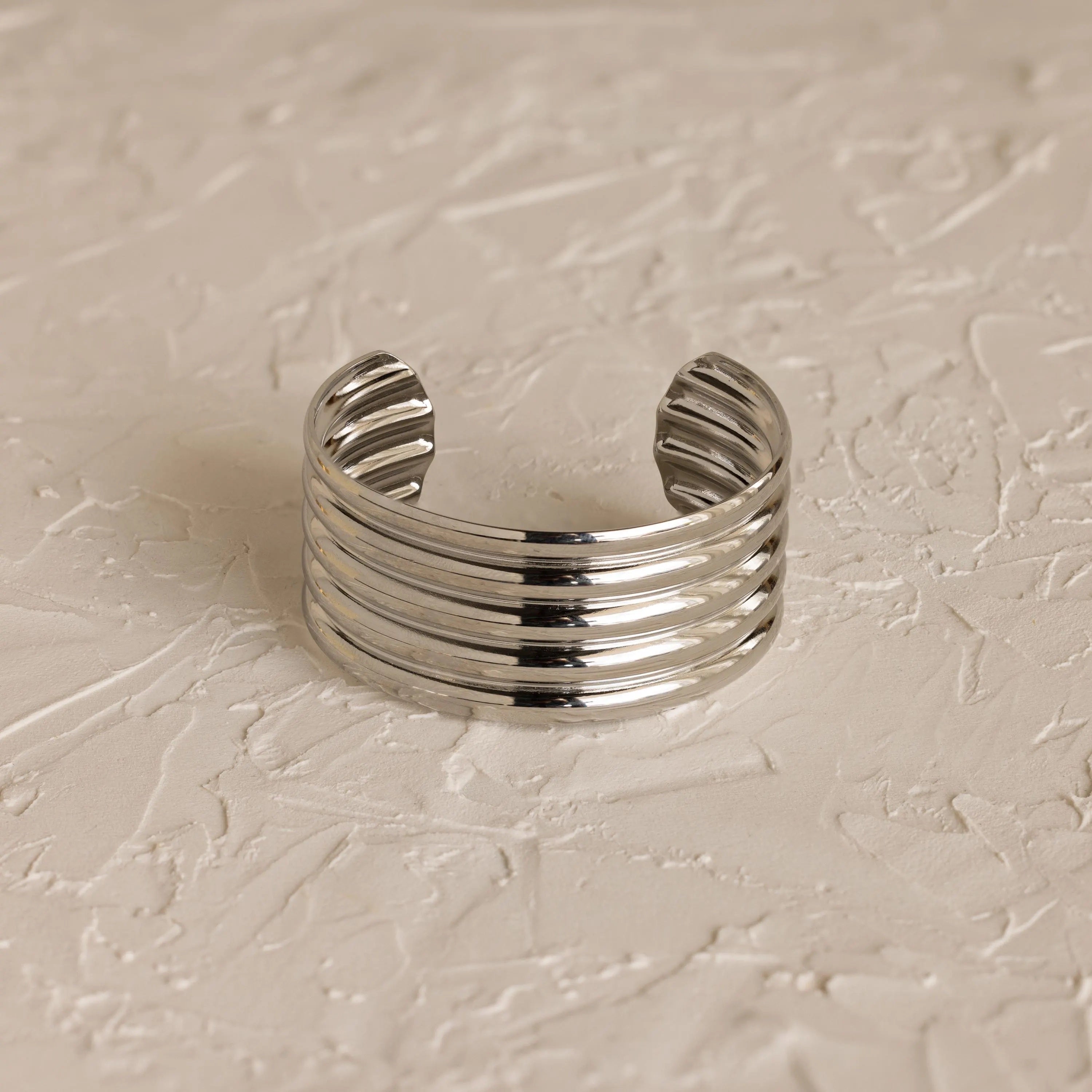The Ribbed Cuff Bracelet is a wide silver sculpted cuff with five raised horizontal ridges, shown on a textured light-colored surface.