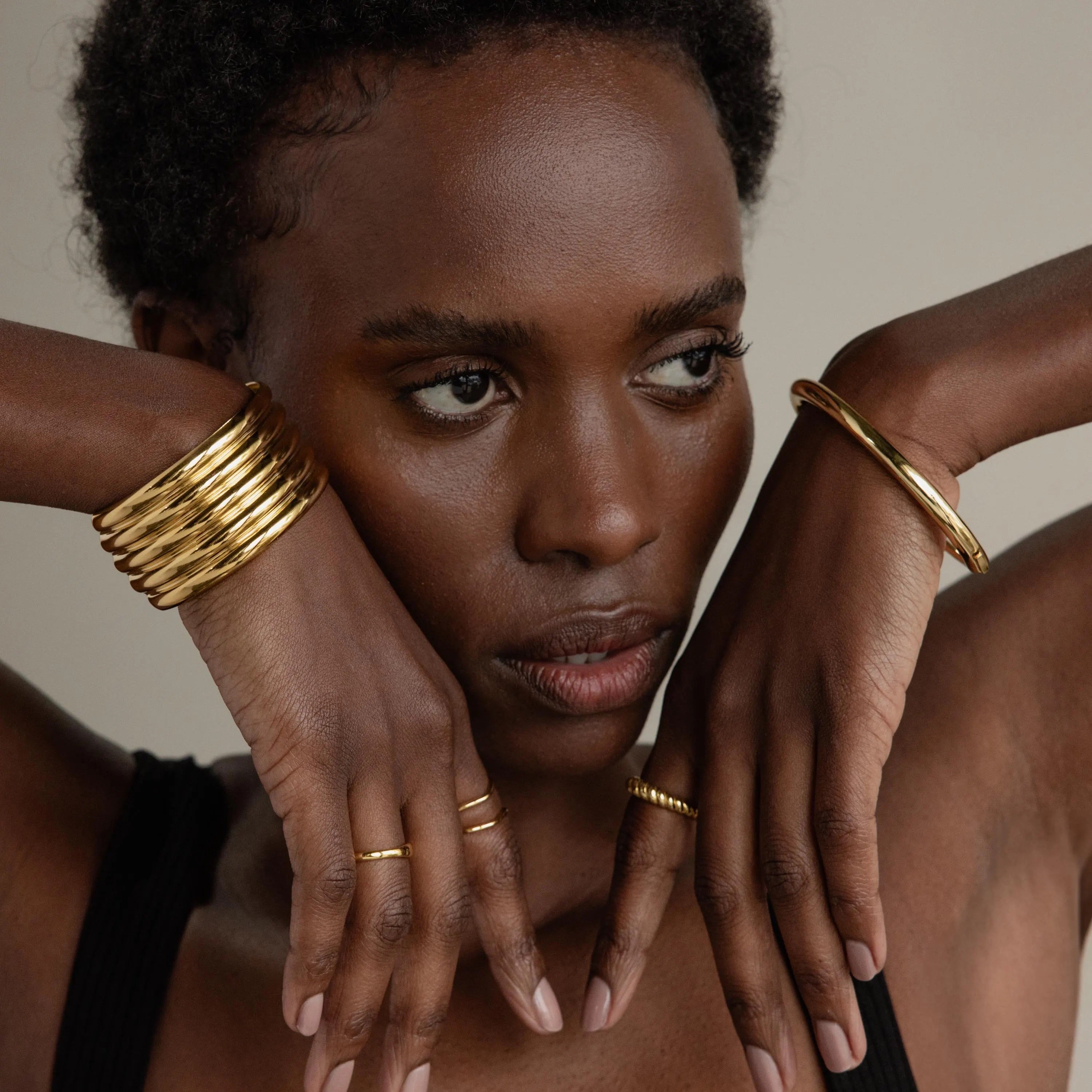 A woman with short hair poses with her hands near her face, wearing gold rings and the Ribbed Cuff Bracelet, set against a neutral background.