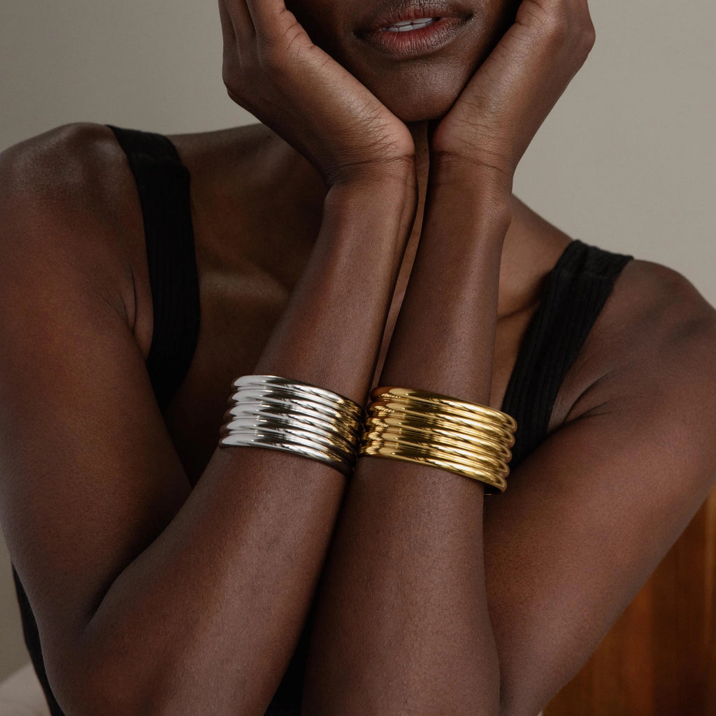 A woman wears chunky gold and silver bangles, including the Ribbed Cuff Bracelet with ridge detailing, as she rests her chin on her crossed hands.