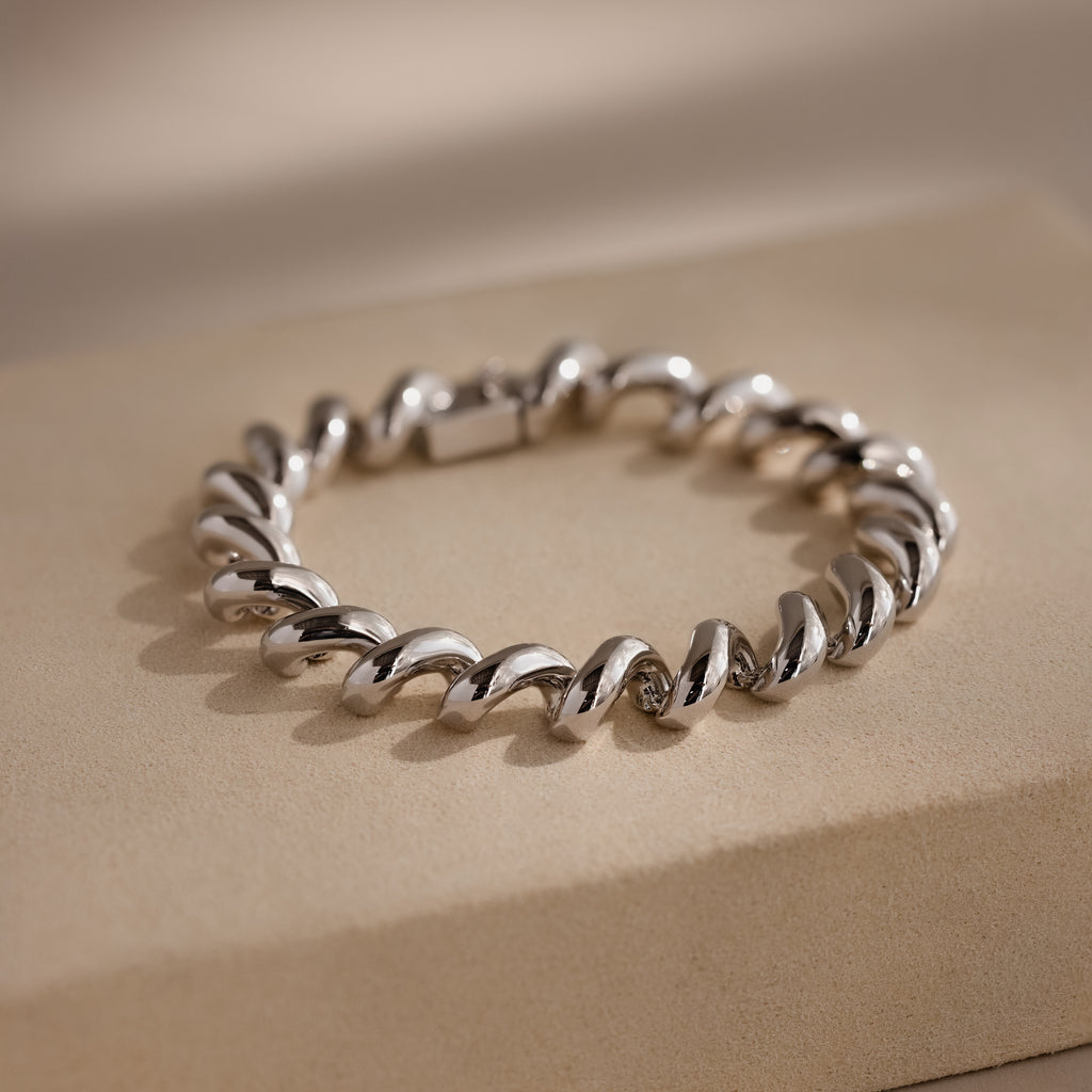 A Twisted Chain Bracelet with bold, gleaming links is displayed on a beige rectangular surface, exuding effortless glamour.