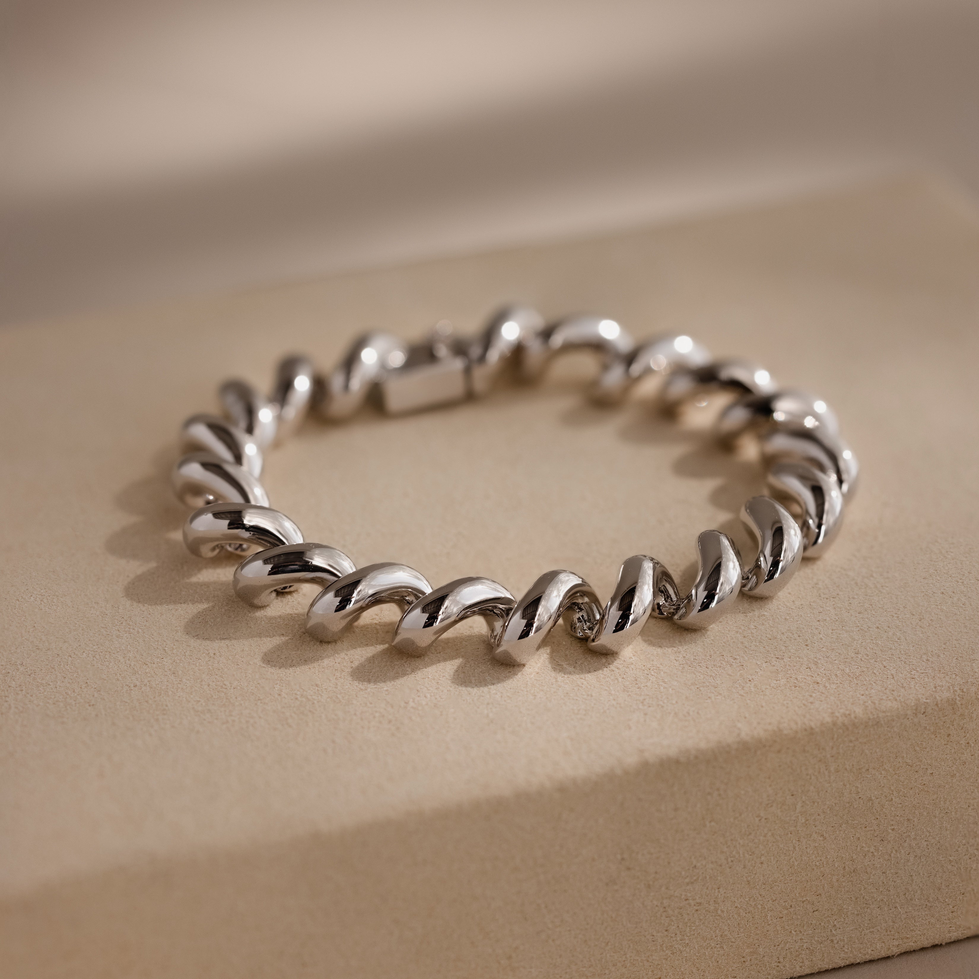 A Twisted Chain Bracelet with bold, gleaming links is displayed on a beige rectangular surface, exuding effortless glamour.