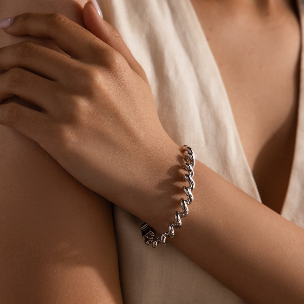 A person wearing a bold Twisted Chain Bracelet in White Gold with shining links rests their hand on their arm, dressed in a light sleeveless top, radiating effortless glamour.