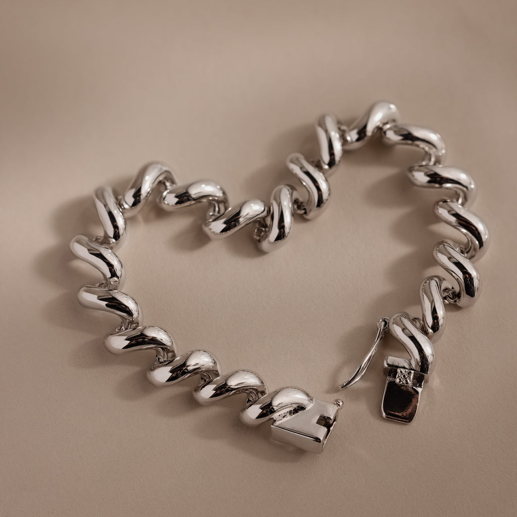 A Twisted Chain Bracelet in White Gold with bold, shining links is shaped into a heart on a beige surface, radiating effortless glamour.