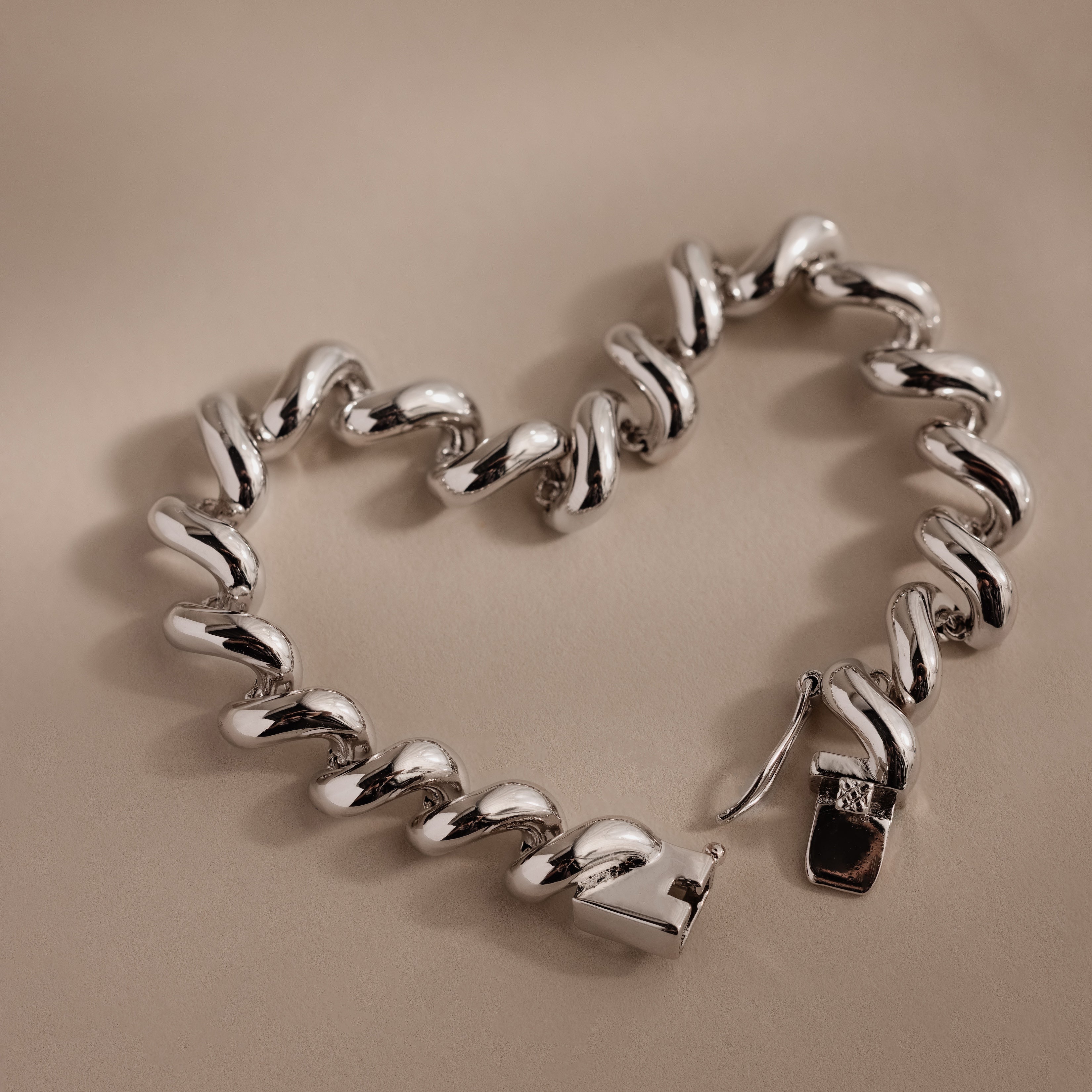 A Twisted Chain Bracelet in White Gold with bold, shining links is shaped into a heart on a beige surface, radiating effortless glamour.