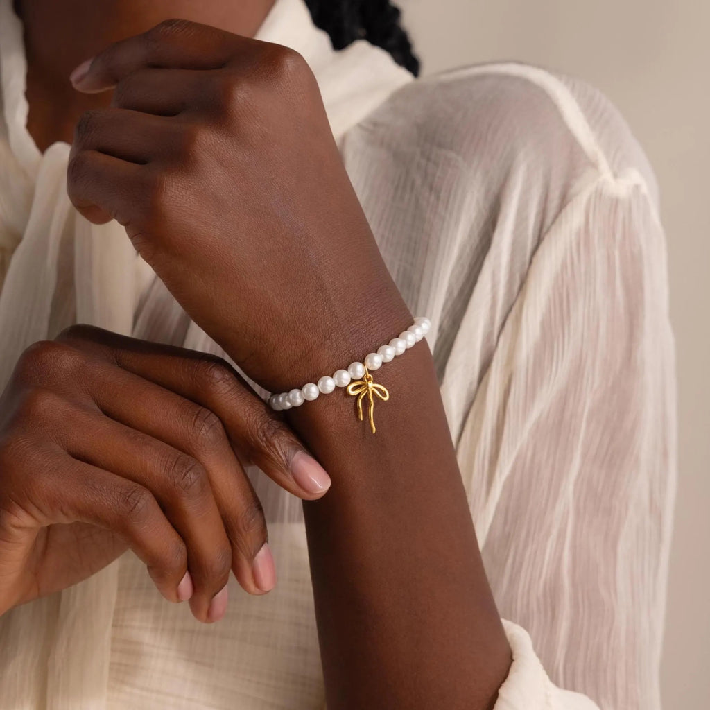 A close-up of a woman’s wrist adorned with the Bow Pearl Bracelet in gold. The bracelet features a strand of lustrous white pearls strung together with a petite, high-polish gold bow charm hanging delicately in the center.