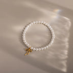 A delicate pearl bracelet arranged in a perfect circle on a soft beige surface. The pearls are smooth and glossy, centered by a dainty yellow gold bow charm that hangs slightly off-center, casting a gentle shadow in the light.