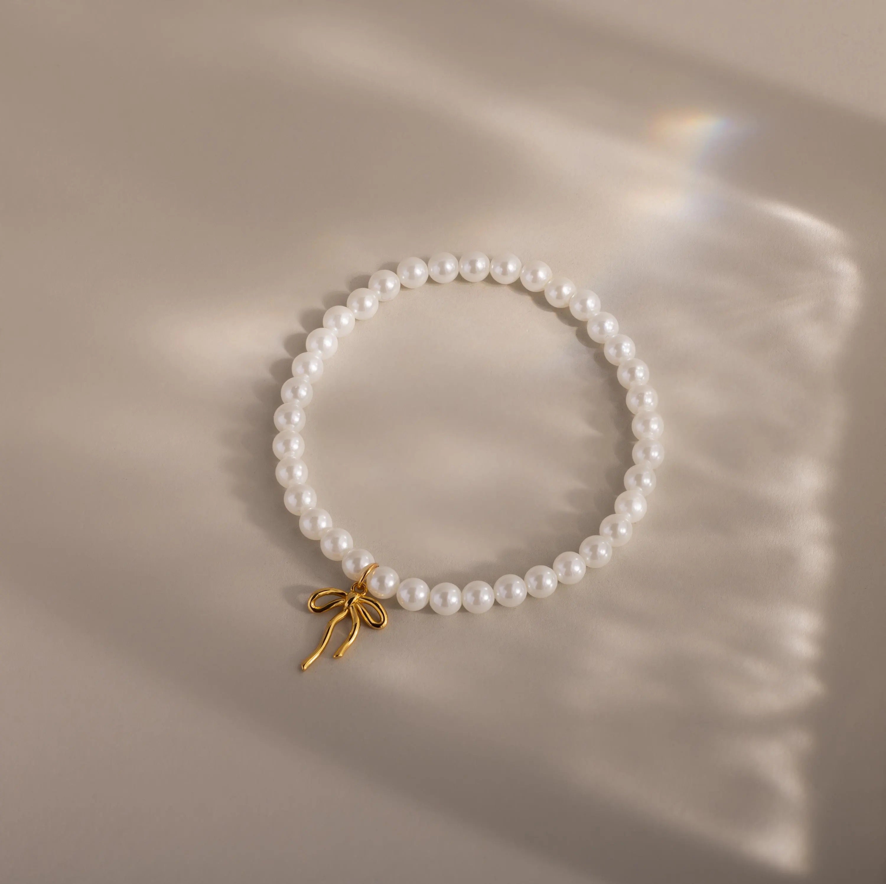 A delicate pearl bracelet arranged in a perfect circle on a soft beige surface. The pearls are smooth and glossy, centered by a dainty yellow gold bow charm that hangs slightly off-center, casting a gentle shadow in the light.