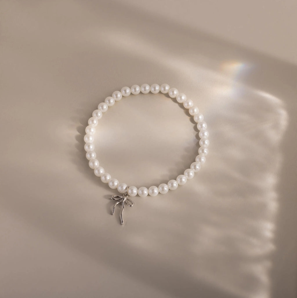 An elegant pearl bracelet with a minimalist white gold bow charm is laid out in a neat circle on a softly illuminated neutral surface.