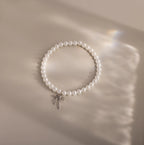 An elegant pearl bracelet with a minimalist white gold bow charm is laid out in a neat circle on a softly illuminated neutral surface.
