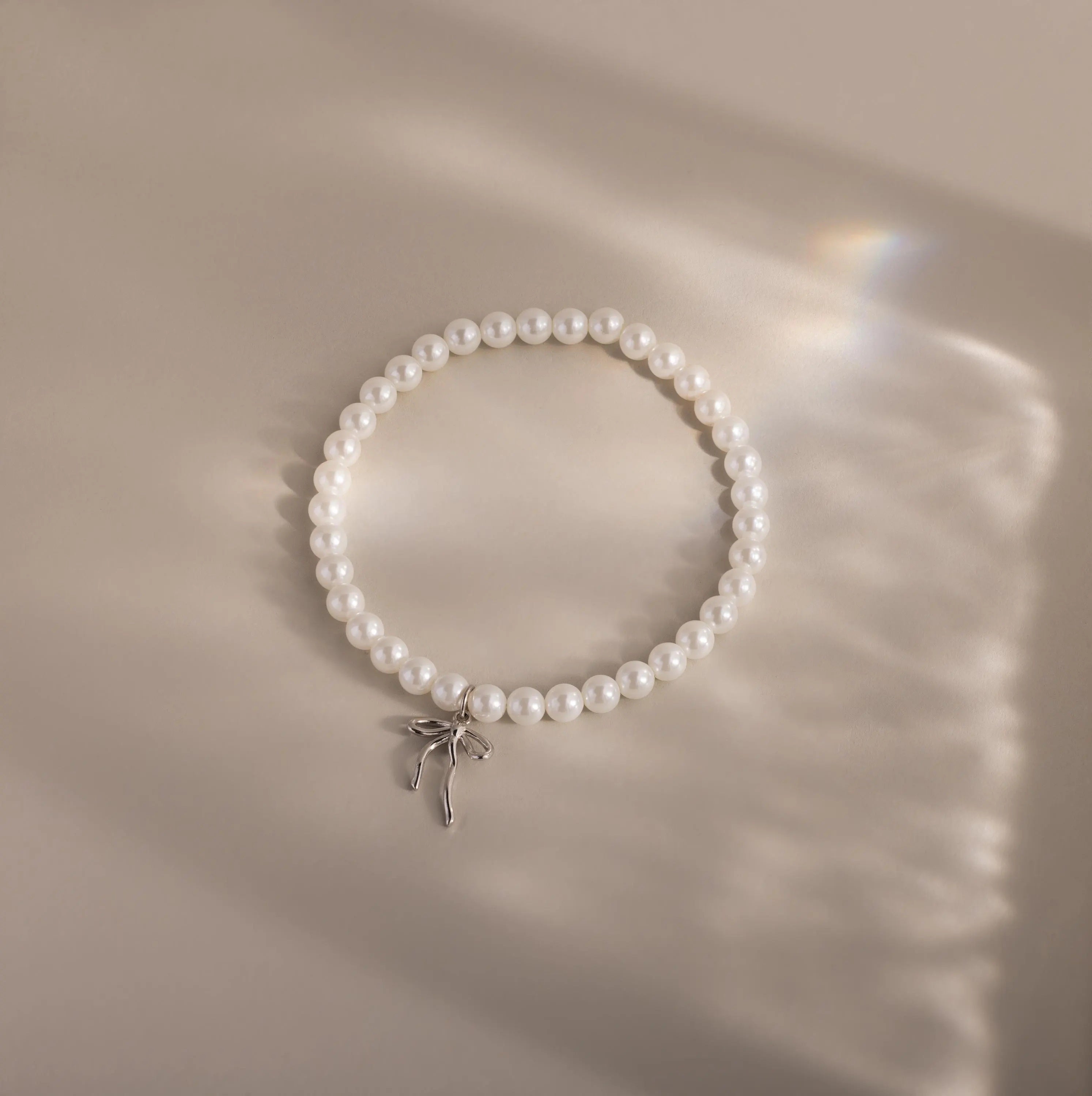 An elegant pearl bracelet with a minimalist white gold bow charm is laid out in a neat circle on a softly illuminated neutral surface.