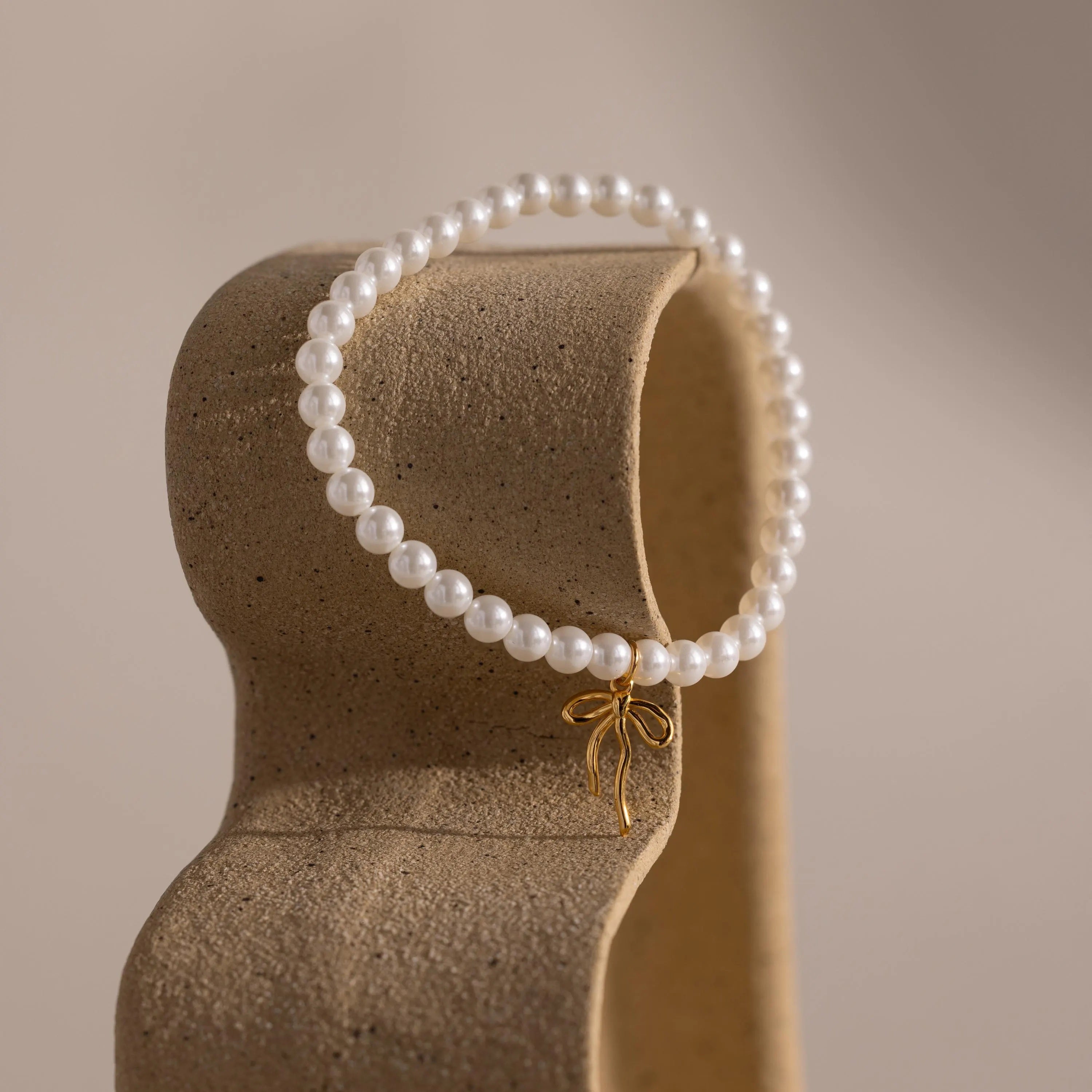 The Bow Pearl Bracelet in gold is draped over a sculptural, curved stand.