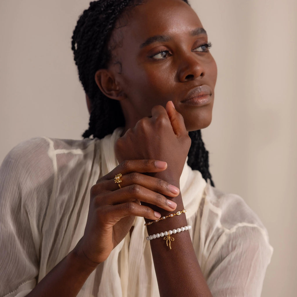 A portrait of a model gazing thoughtfully into the distance, wearing the Bow Pearl Bracelet in gold stacked with another bracelet and gold ring.