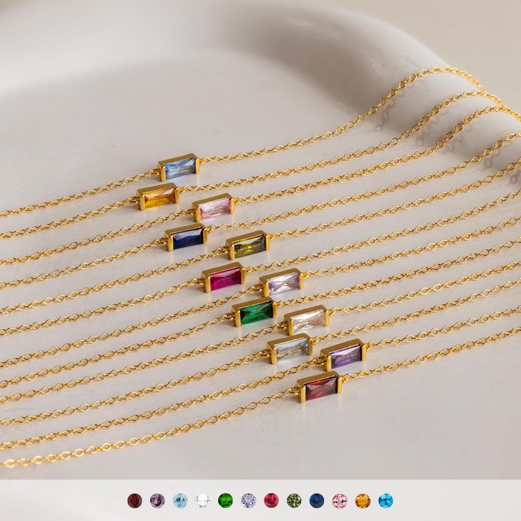 Ten vibrant Dainty Baguette Birthstone Bracelets with colorful rectangular charms are lined up on a light surface—each minimalist piece is perfect for stacking.
