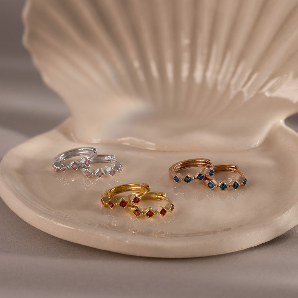 Four Geometric Birthstone Huggies with diamond accents rest on a ceramic shell dish, showcasing gold, silver, and rose gold tones—a perfect touch of birthstone jewelry elegance.