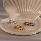 Four Geometric Birthstone Huggies with diamond accents rest on a ceramic shell dish, showcasing gold, silver, and rose gold tones—a perfect touch of birthstone jewelry elegance.