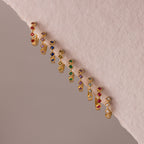 The Geometric Alexandrite Huggies are gold earrings with colorful gemstones in rows and subtle diamond accents, displayed on a light pink textured surface.