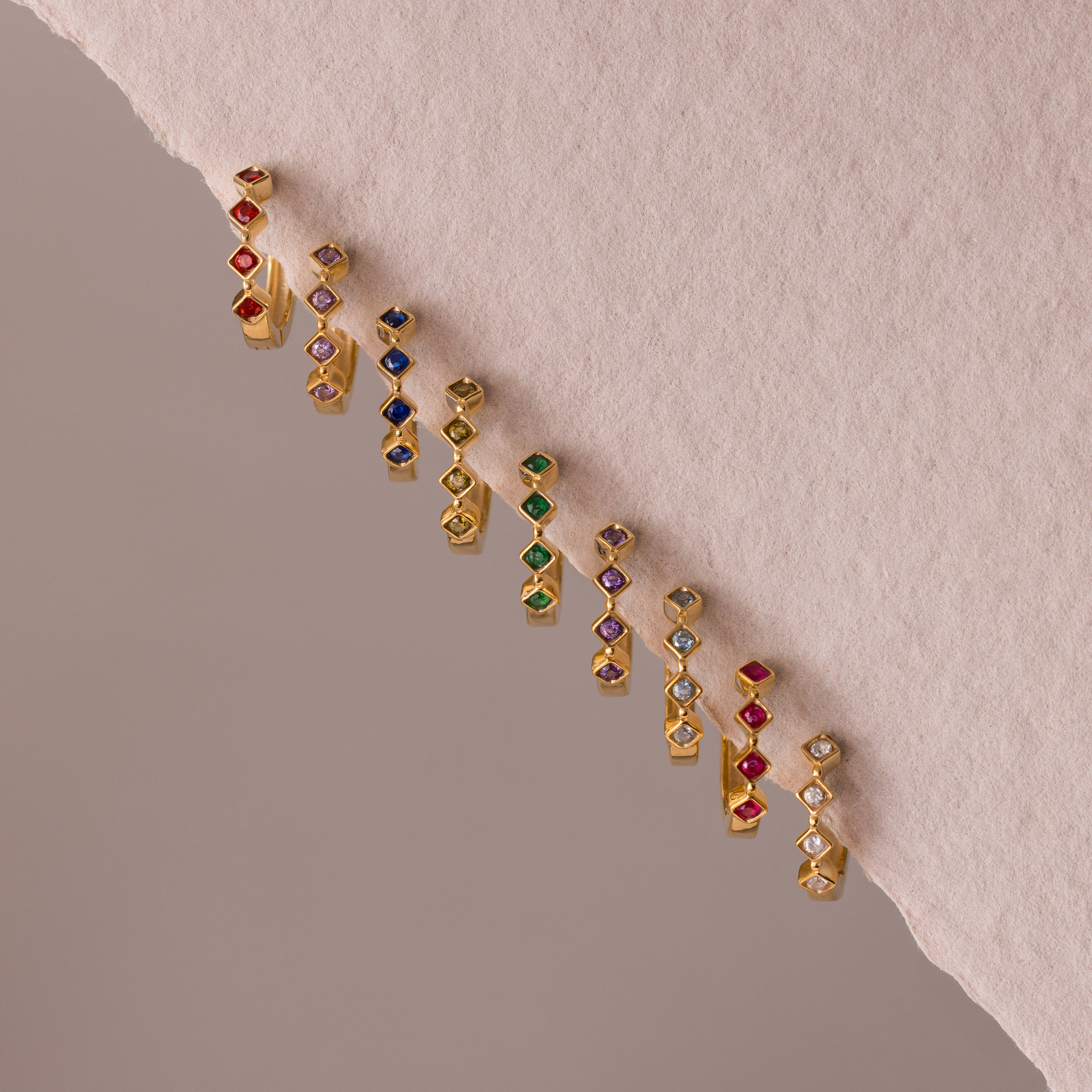 The Geometric Alexandrite Huggies are gold earrings with colorful gemstones in rows and subtle diamond accents, displayed on a light pink textured surface.
