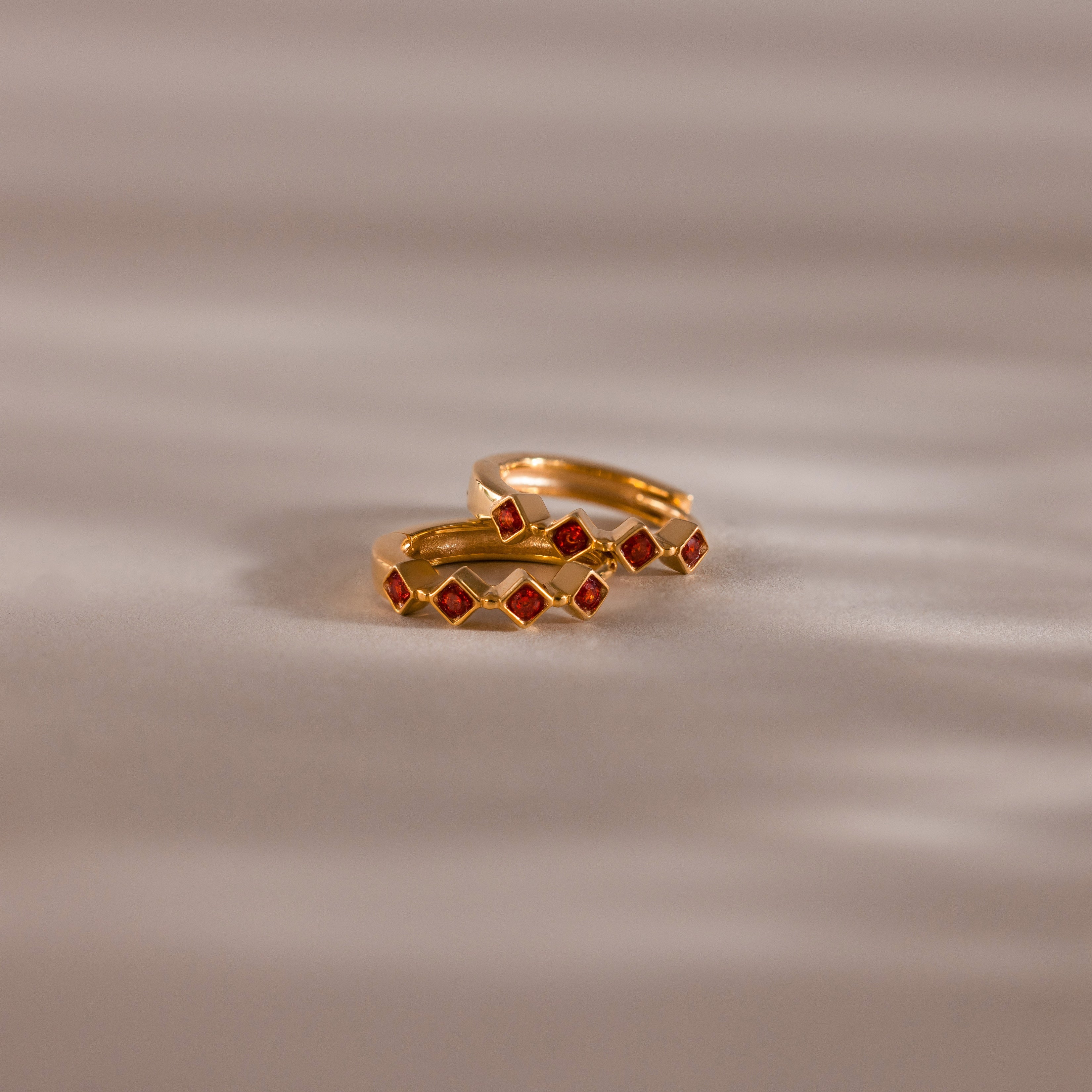 Geometric Garnet Huggies: Two gold rings with small red square garnet stones and diamond accents rest on a softly lit beige surface, offering a modern twist on birthstone jewelry.
