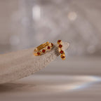 Geometric Garnet Huggies—gold earrings featuring small red garnet stones, reminiscent of birthstone jewelry—displayed on a textured, light-colored stone surface.