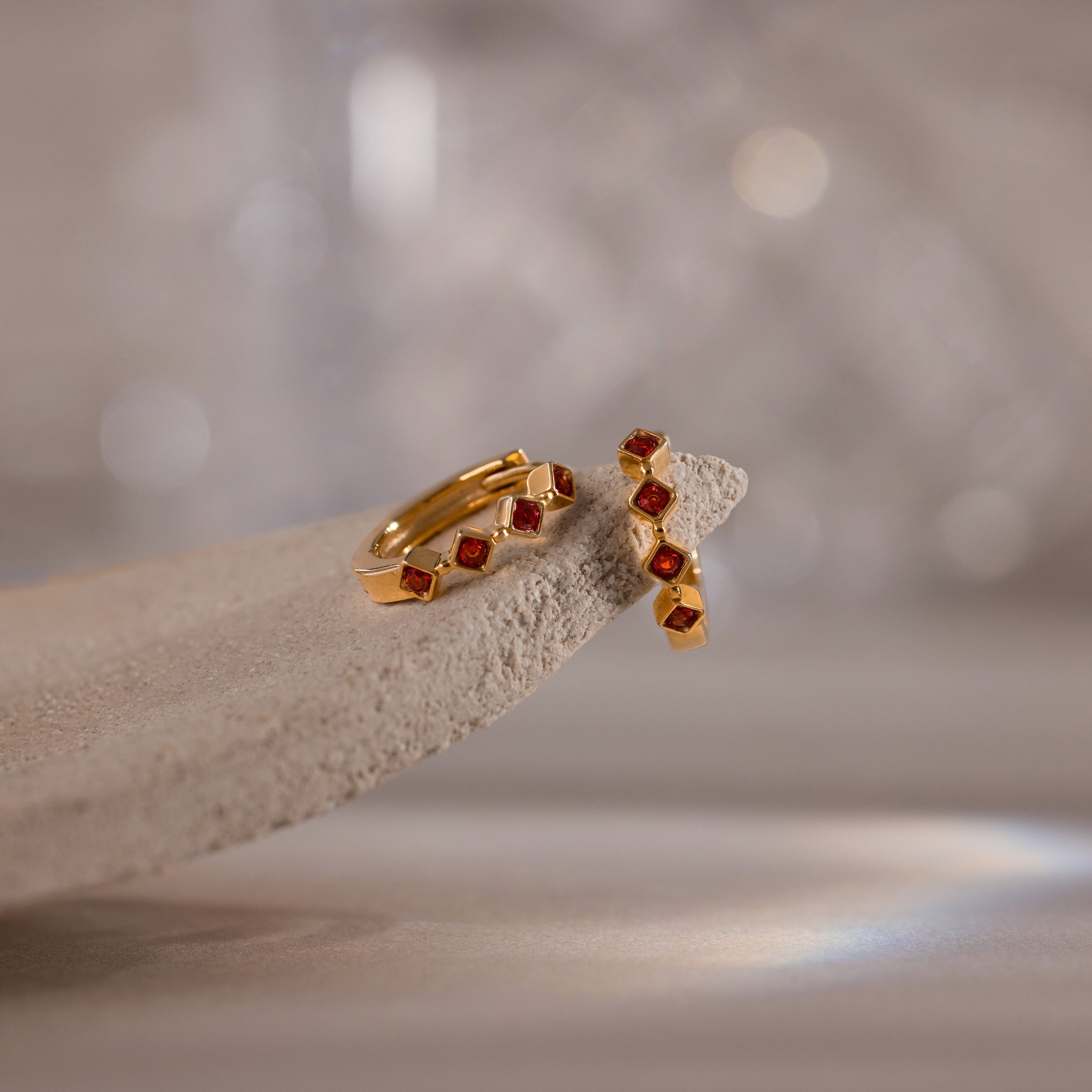 Geometric Garnet Huggies—gold earrings featuring small red garnet stones, reminiscent of birthstone jewelry—displayed on a textured, light-colored stone surface.
