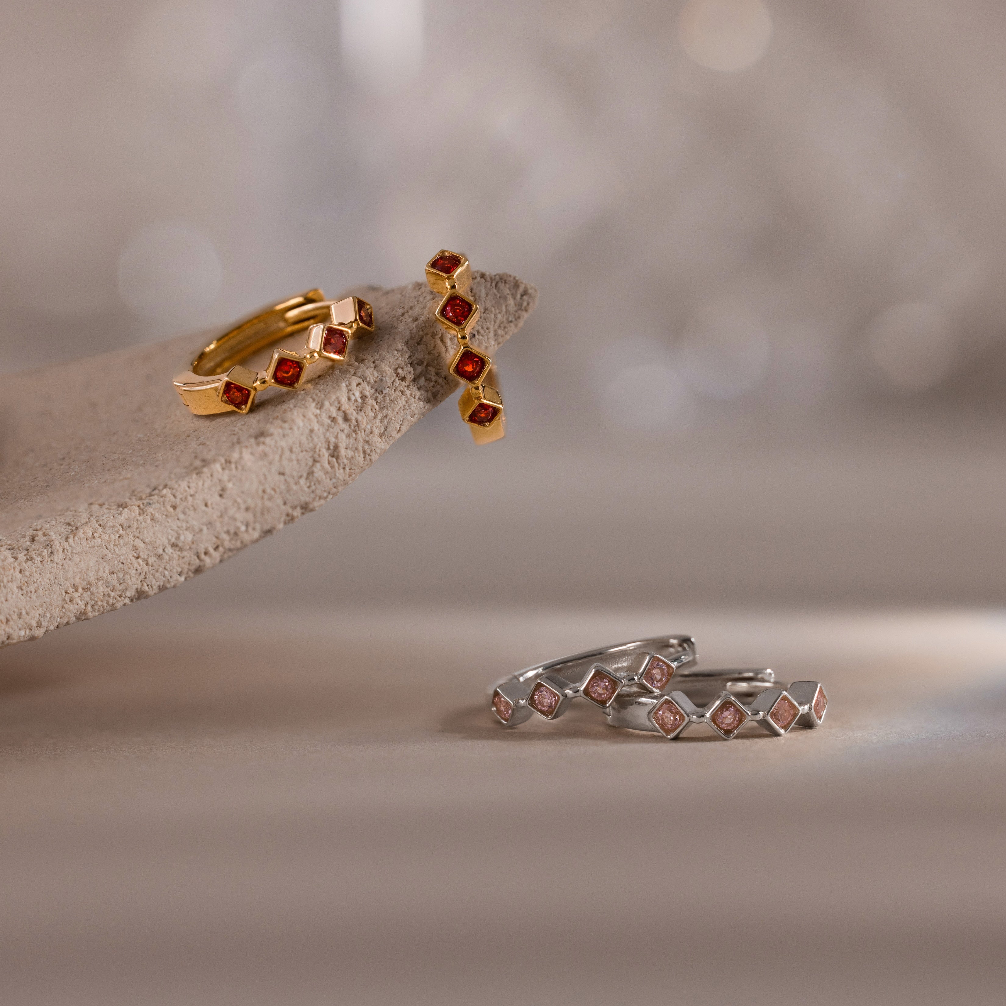 Two pairs of Geometric Birthstone Huggies—one gold, one silver—feature small red stones and subtle diamond accents, blending modern style with elegant design.