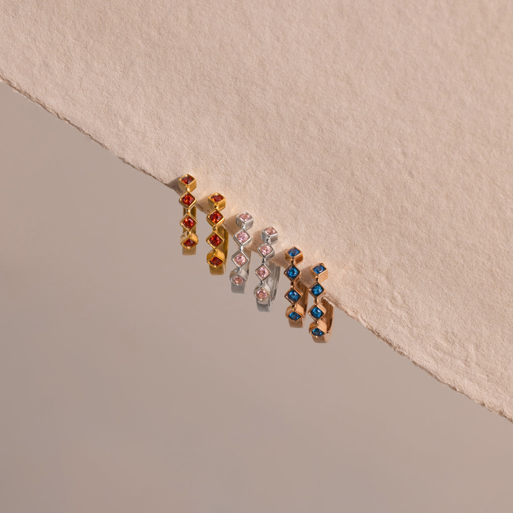 The Geometric Blue Zircon Huggies add a vibrant touch to your jewelry collection with their striking blue stones, shown against a beige textured background.