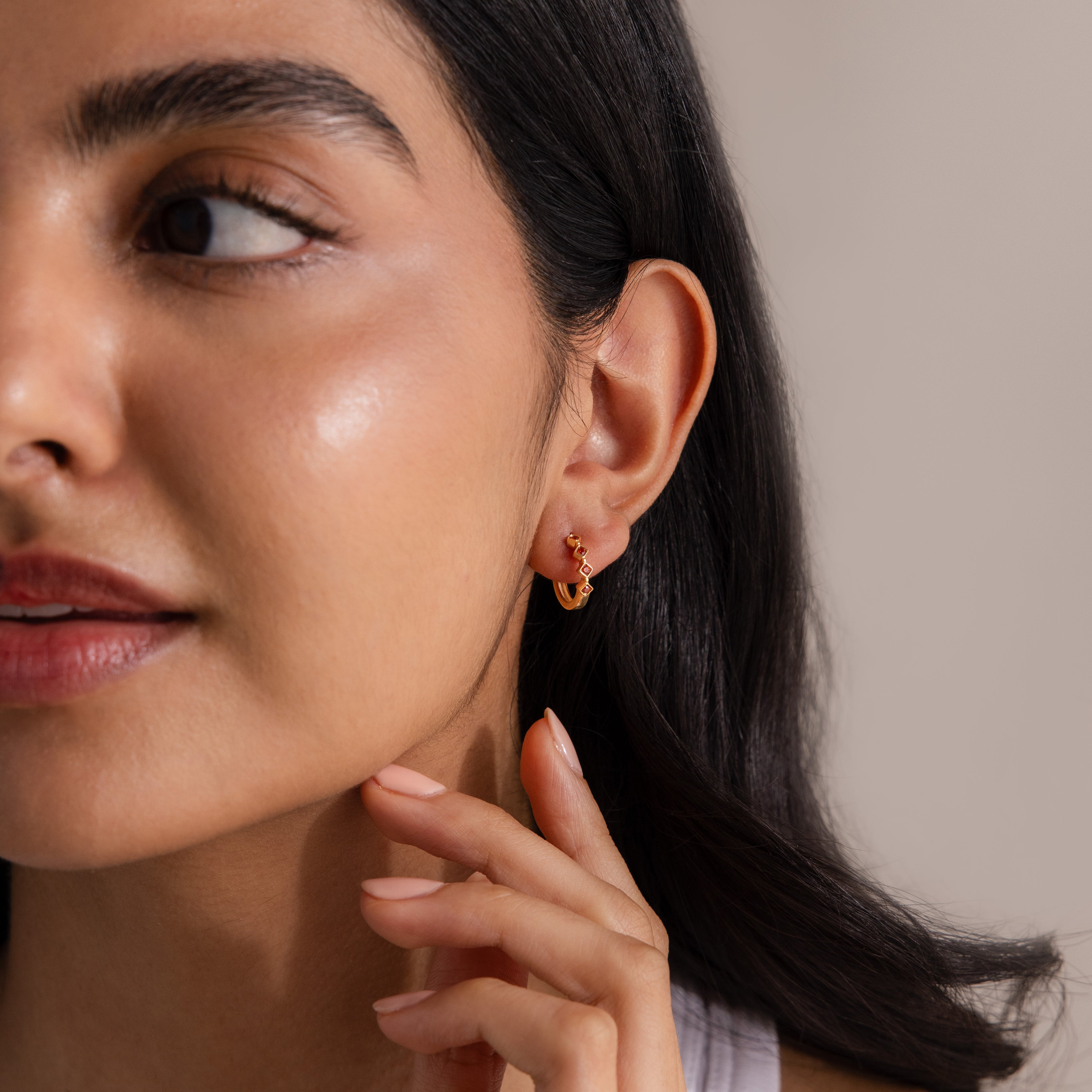 Woman with smooth skin wearing Geometric Onyx Huggies—gold hoops accented with delicate diamonds—touches her face under soft lighting and a neutral background.