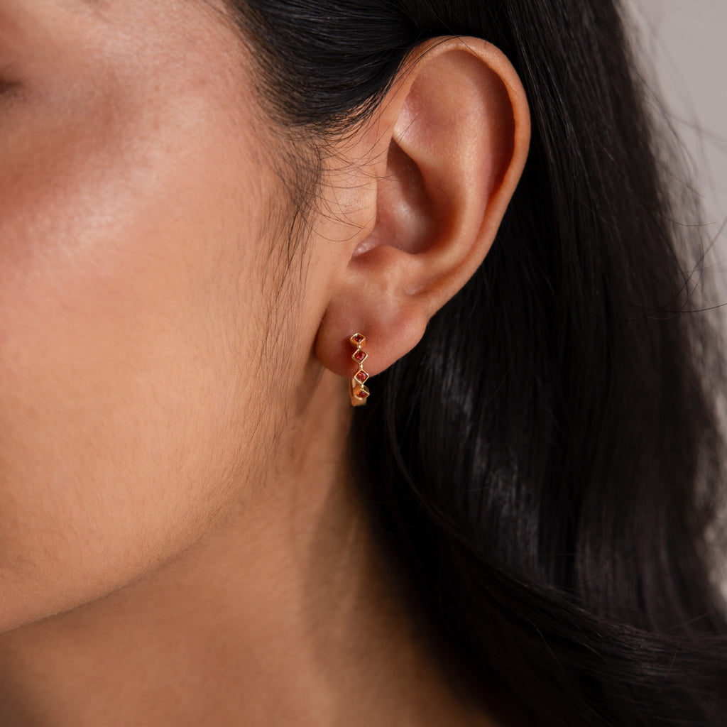 Close-up of a woman’s ear wearing Geometric Onyx Huggies, small gold hoop earrings accented with black onyx; her straight dark hair is visible.