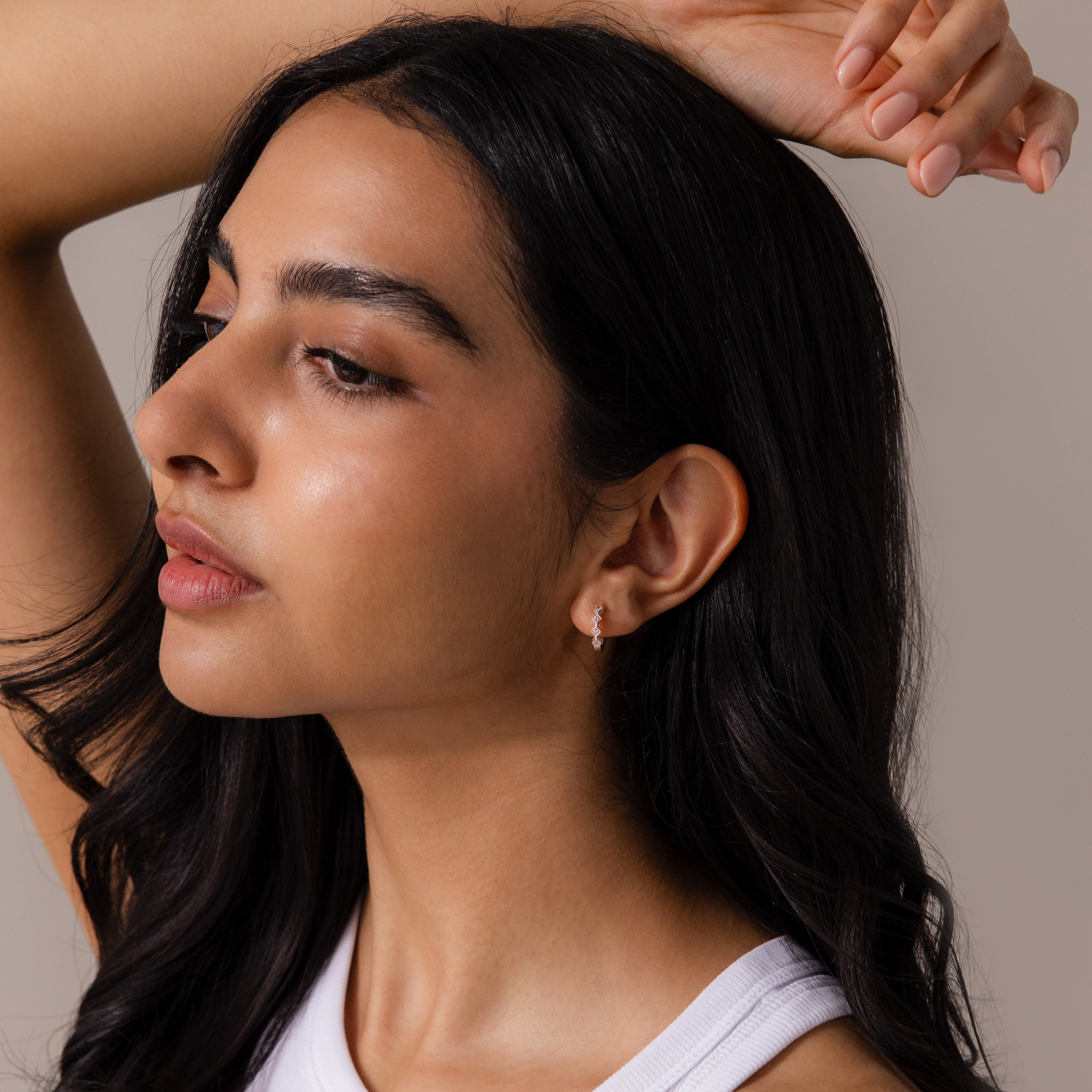 Woman with long dark hair and glowing skin poses with arm raised, wearing a white top and the Geometric Pink Tourmaline Huggies sparkling in her ears.