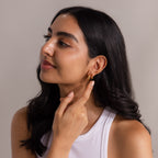 Woman with long dark hair in a white tank top touches her Geometric Topaz Huggies, gold hoop earrings with sparkling accents, while smiling slightly.