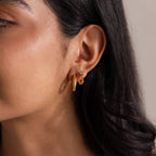 Close-up of a woman's ear adorned with gold hoops and Geometric Alexandrite Huggies, her long dark hair elegantly framing the birthstone earrings.