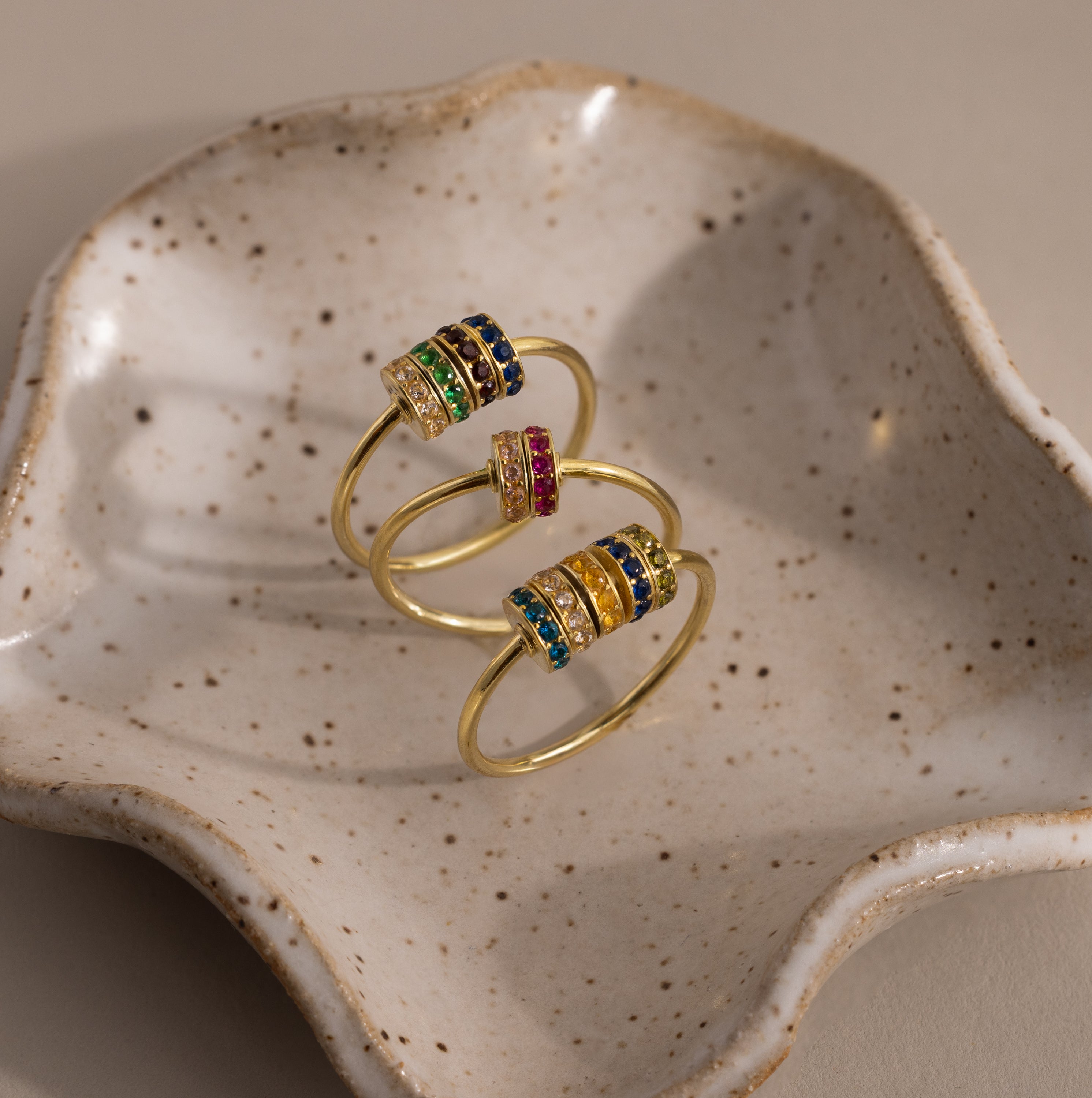 The Multiple Birthstone Bead Ring in 18K Gold features colorful gemstone bands and rests on a speckled, shell-shaped ceramic dish—an ideal pick for lovers of personalized jewelry.
